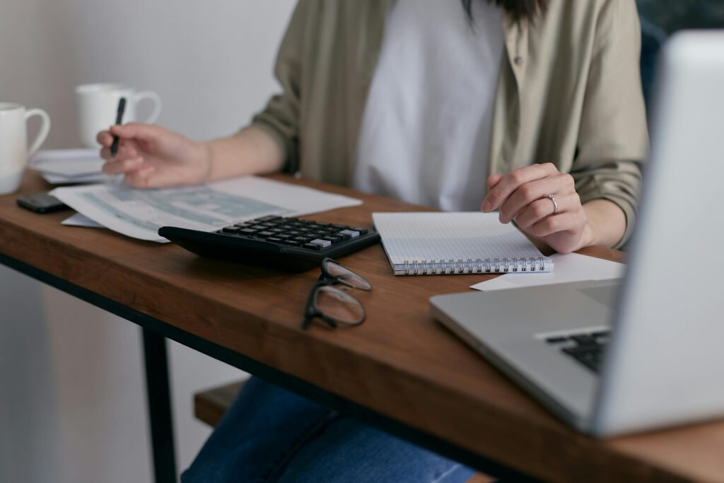 a person writing on a table with paper, calculator and laptop